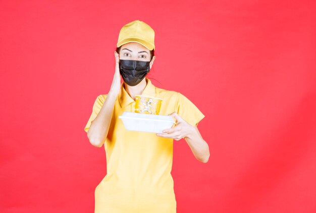 Mensajero femenino en uniforme amarillo y máscara negra sosteniendo un paquete de comida para llevar y parece confundido y pensativo