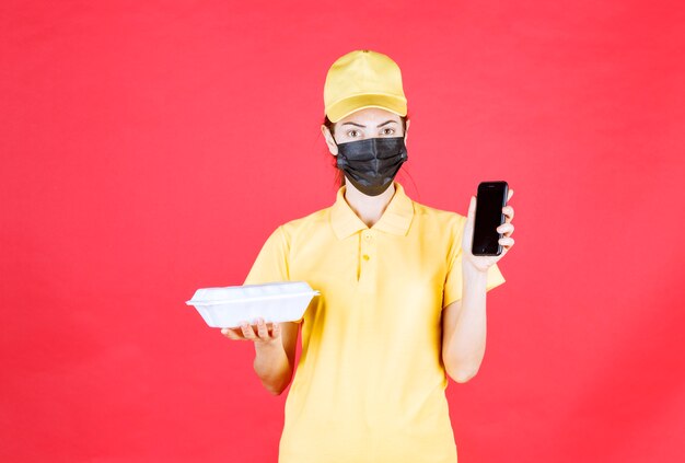 Mensajero femenino en uniforme amarillo y máscara negra sosteniendo un paquete de comida para llevar y tomando pedidos a través de un teléfono inteligente