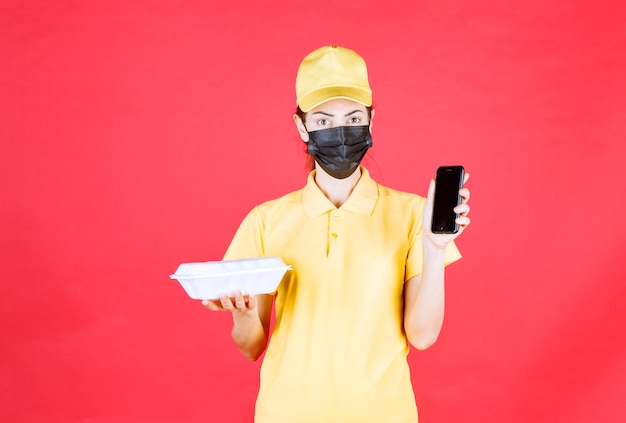 Mensajero femenino en uniforme amarillo y máscara negra sosteniendo un paquete de comida para llevar y tomando pedidos a través de un teléfono inteligente
