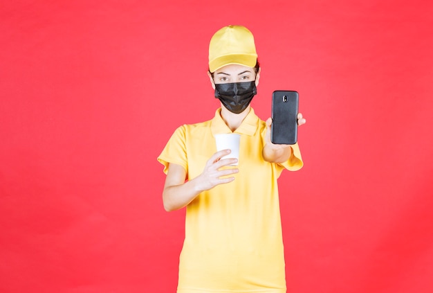 Mensajero femenino en uniforme amarillo y máscara negra sosteniendo una taza para llevar y hablando por teléfono o tomando pedidos