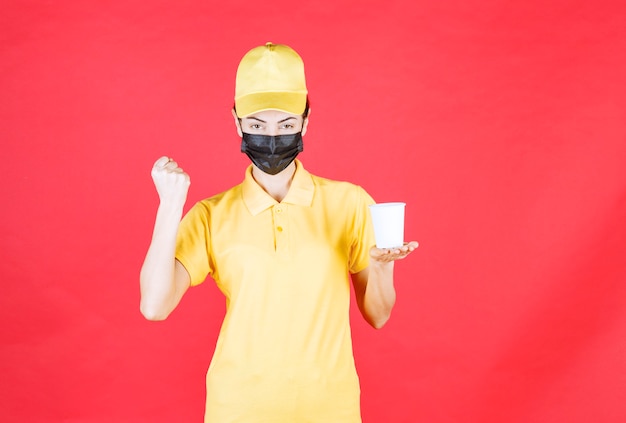 Mensajero femenino en uniforme amarillo y máscara negra sosteniendo una taza para llevar y mostrando su puño