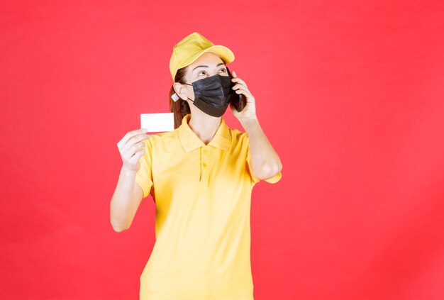 Mensajero femenino en uniforme amarillo y máscara negra sosteniendo un teléfono inteligente y presentando su tarjeta de visita mientras habla por teléfono