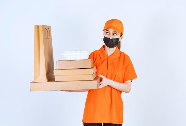 Mensajero femenino en uniforme amarillo y máscara negra sosteniendo varios paquetes de cartón, caja de comida para llevar y bolsa de compras mientras se ve confundido y pensativo.