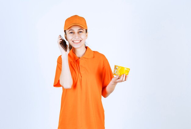 Mensajero femenino en uniforme amarillo sosteniendo una taza de fideos para llevar y tomando el pedido desde el teléfono inteligente