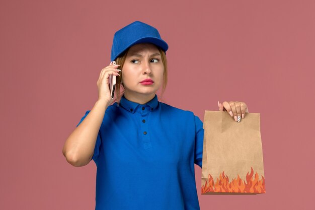 Mensajero femenino en uniforme azul hablando por teléfono sosteniendo el paquete de alimentos en rosa claro, trabajo de entrega uniforme de servicio