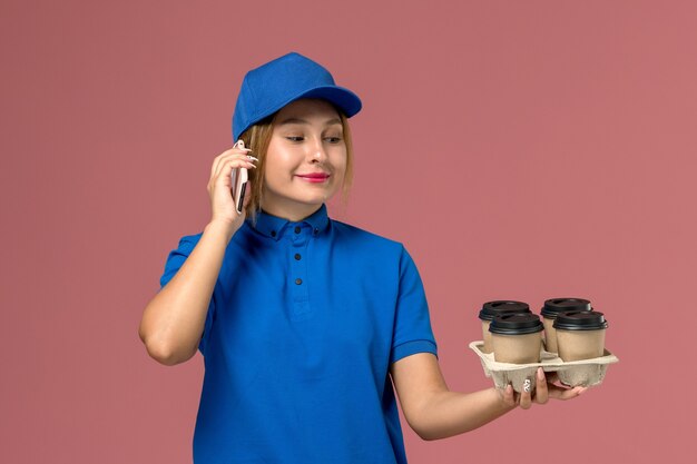 Mensajero femenino en uniforme azul hablando por teléfono sosteniendo tazas de café marrón sonriendo en rosa claro, trabajo de entrega uniforme de servicio