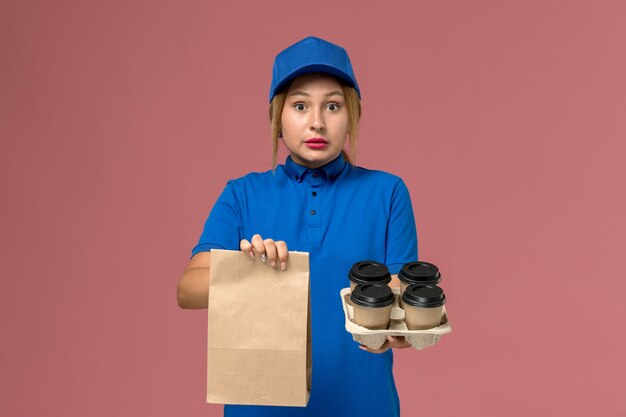 Mensajero femenino en uniforme azul con paquete de comida y tazas de café de entrega marrón en rosa, trabajador de entrega uniforme de servicio