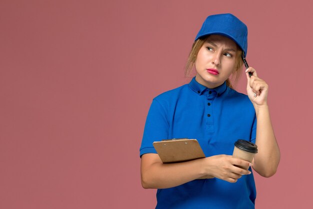 Mensajero femenino en uniforme azul posando sosteniendo una taza de café y un bloc de notas con expresión de pensamiento en rosa, servicio de entrega uniforme