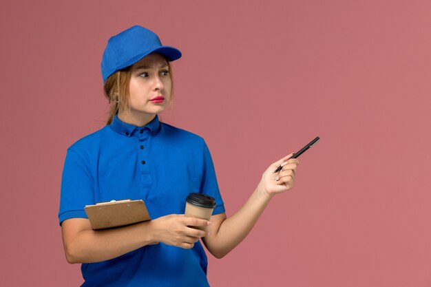 Mensajero femenino en uniforme azul posando sosteniendo una taza de café y un bloc de notas en rosa, servicio uniforme de repartidor trabajadora