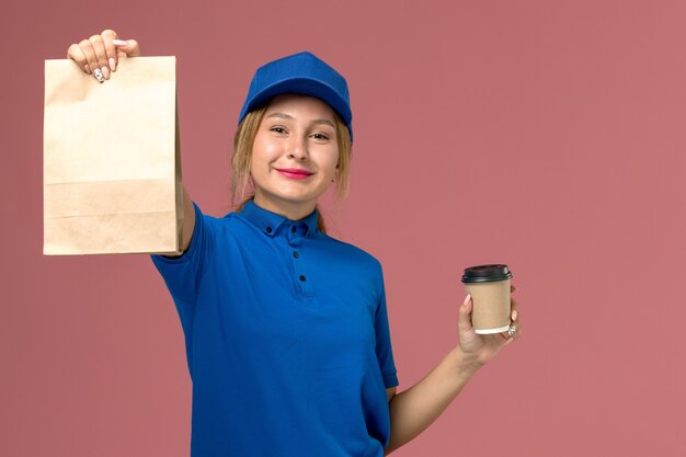 Mensajero femenino en uniforme azul posando sosteniendo una taza de café y paquete de comida con una leve sonrisa en rosa, trabajador de trabajo de entrega uniforme de servicio