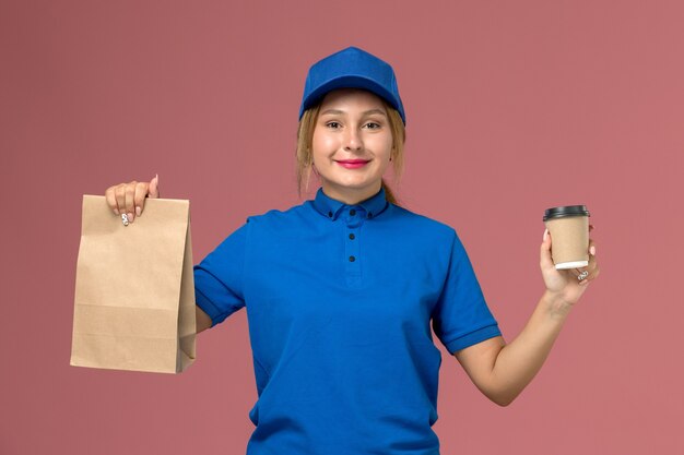 Mensajero femenino en uniforme azul posando sosteniendo una taza de café y paquete de comida en rosa, servicio uniforme de repartidor trabajadora