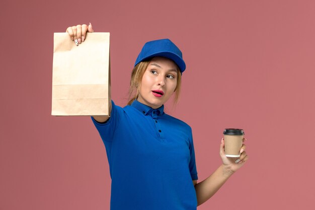 Mensajero femenino en uniforme azul posando sosteniendo una taza de café y paquete de comida en rosa, trabajador de entrega uniforme de servicio