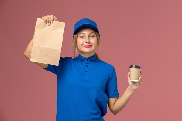 Mensajero femenino en uniforme azul posando sosteniendo una taza de café y paquete de comida sonriendo en rosa, servicio uniforme repartidor trabajadora