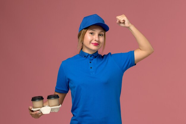 Mensajero femenino en uniforme azul posando y sosteniendo tazas de café flexionando en rosa, trabajo de entrega uniforme de servicio