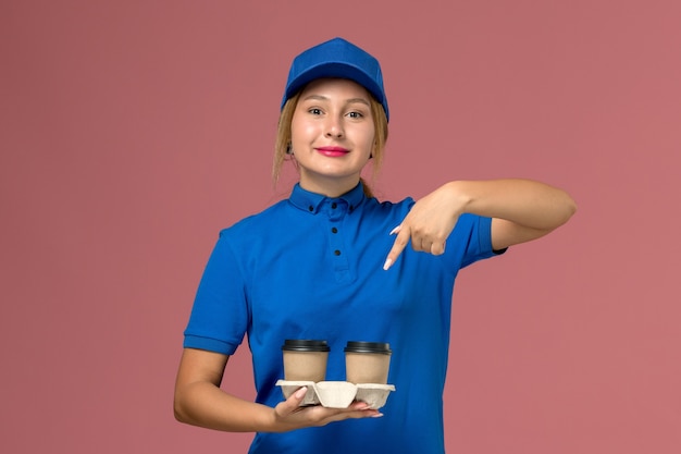 Mensajero femenino en uniforme azul posando y sosteniendo tazas de café con una leve sonrisa en rosa, trabajo de entrega uniforme de servicio