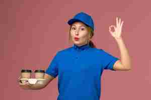 Foto gratuita mensajero femenino en uniforme azul posando y sosteniendo tazas de café mostrando bien firmar en rosa, servicio de entrega uniforme