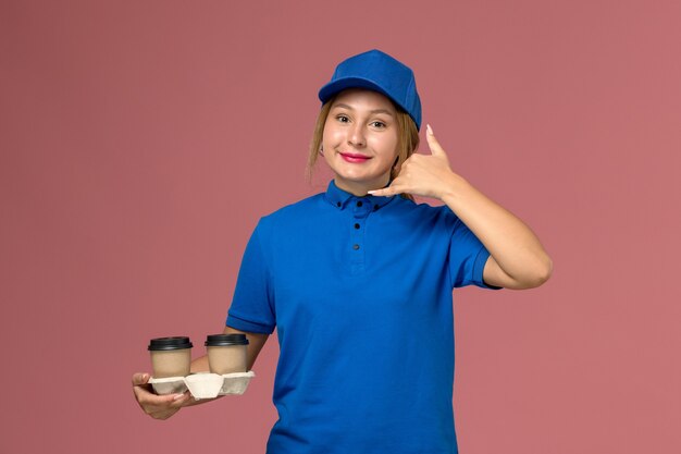 Mensajero femenino en uniforme azul posando y sosteniendo tazas de café mostrando gesto de llamada telefónica en rosa, trabajo de entrega uniforme de servicio