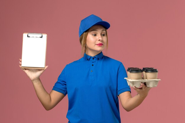 Mensajero femenino en uniforme azul sosteniendo el bloc de notas junto con tazas de café de entrega marrón en rosa claro, servicio de trabajo entrega uniforme