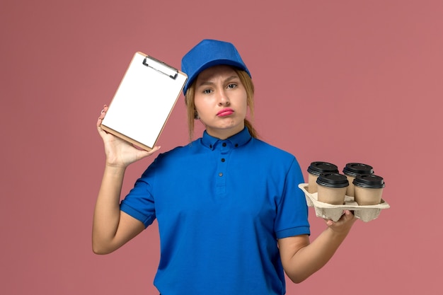 Mensajero femenino en uniforme azul sosteniendo el bloc de notas junto con tazas de café de entrega marrón en rosa claro, servicio de trabajo entrega uniforme