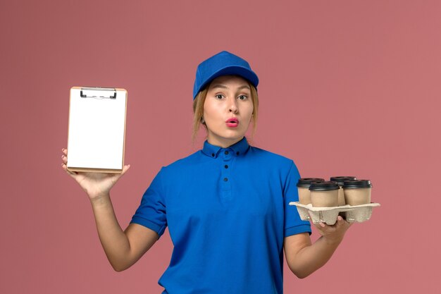 Mensajero femenino en uniforme azul sosteniendo el bloc de notas junto con tazas de café de entrega marrón en rosa claro, trabajo de entrega uniforme de servicio