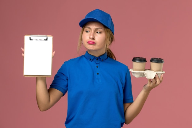 Mensajero femenino en uniforme azul sosteniendo tazas de café y bloc de notas pensando en rosa, servicio de entrega uniforme del trabajador