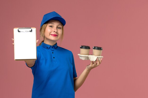 Mensajero femenino en uniforme azul sosteniendo tazas de café y bloc de notas en rosa, servicio de entrega uniforme del trabajador