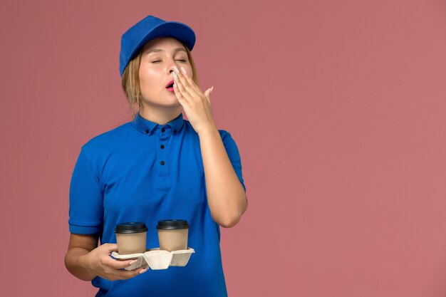 Mensajero femenino en uniforme azul sosteniendo tazas de café bostezando en rosa, trabajo de entrega uniforme de servicio