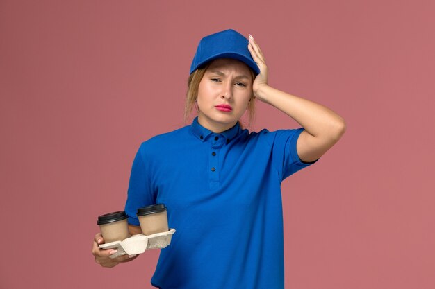 Mensajero femenino en uniforme azul sosteniendo tazas de café con dolor de cabeza en rosa, trabajo de entrega uniforme de servicio