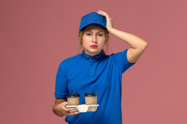 Mensajero femenino en uniforme azul sosteniendo tazas de café con dolor de cabeza en rosa, trabajo de entrega uniforme de servicio