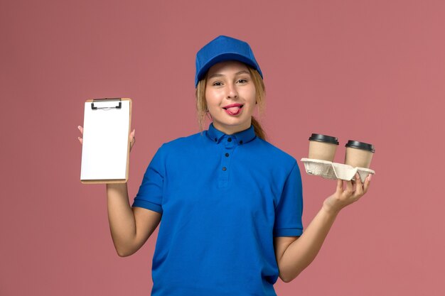 Mensajero femenino en uniforme azul sosteniendo tazas de café de entrega y bloc de notas con lengua en rosa, entrega uniforme del trabajador de servicio