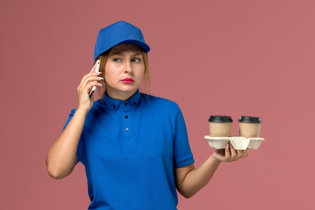 Mensajero femenino en uniforme azul sosteniendo tazas de café de entrega y hablando por teléfono en rosa, servicio de entrega uniforme del trabajador