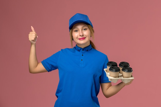 Mensajero femenino en uniforme azul sosteniendo tazas de café de entrega marrón con una leve sonrisa en rosa, trabajador de trabajo de entrega uniforme de servicio