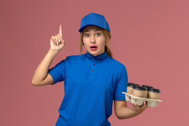 Mensajero femenino en uniforme azul sosteniendo tazas de café de entrega marrón posando con el dedo levantado en rosa, servicio de entrega uniforme