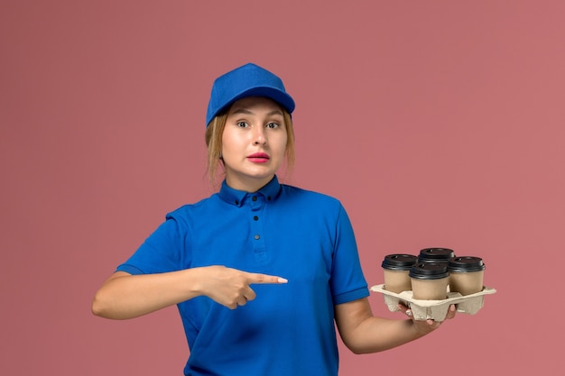 Mensajero femenino en uniforme azul sosteniendo tazas de café de entrega marrón señalando en rosa, trabajador de trabajo de entrega uniforme de servicio
