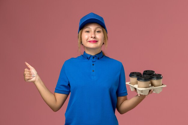 Mensajero femenino en uniforme azul sosteniendo tazas de café de entrega mostrando como signo en rosa, entrega uniforme del trabajador de servicio