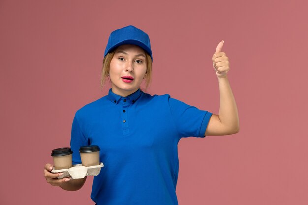 Mensajero femenino en uniforme azul sosteniendo tazas de café de entrega mostrando como signo en rosa, trabajo de entrega uniforme de servicio