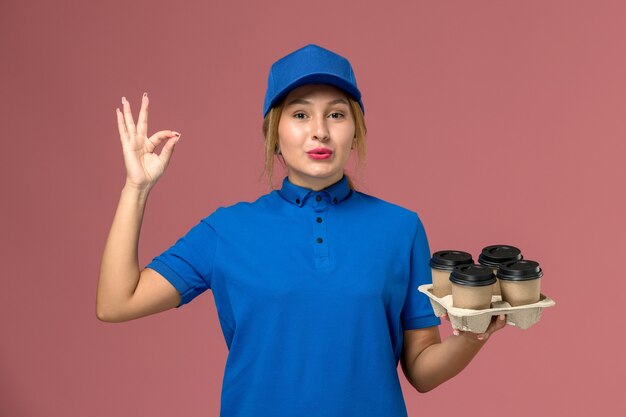 Mensajero femenino en uniforme azul sosteniendo tazas de café de entrega posando en rosa, entrega uniforme del trabajador de servicio