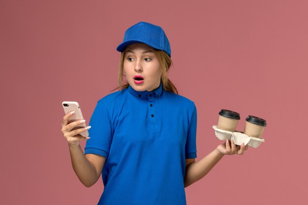 Mensajero femenino en uniforme azul sosteniendo tazas de café de entrega y usando su teléfono con expresión de sorpresa en rosa, servicio de entrega uniforme