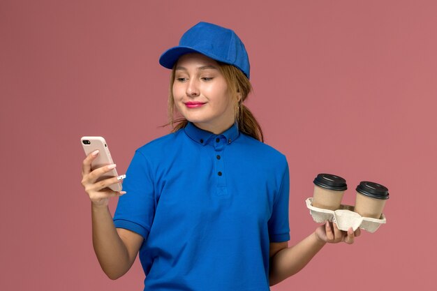 Mensajero femenino en uniforme azul sosteniendo tazas de café de entrega y usando su teléfono en rosa, servicio de entrega uniforme del trabajador