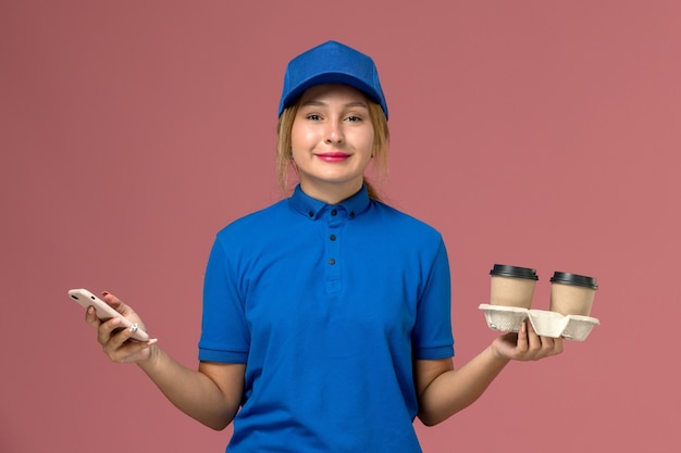 Mensajero femenino en uniforme azul sosteniendo tazas de café de entrega y usando su teléfono en rosa, trabajo de entrega uniforme del trabajador de servicio