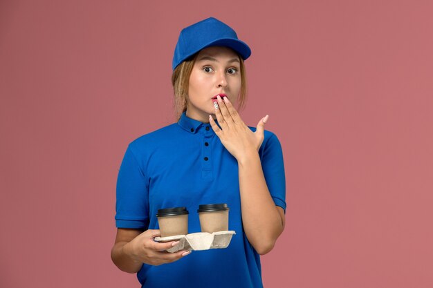 Mensajero femenino en uniforme azul sosteniendo tazas de café con expresión de sorpresa en rosa, trabajo de entrega uniforme de servicio