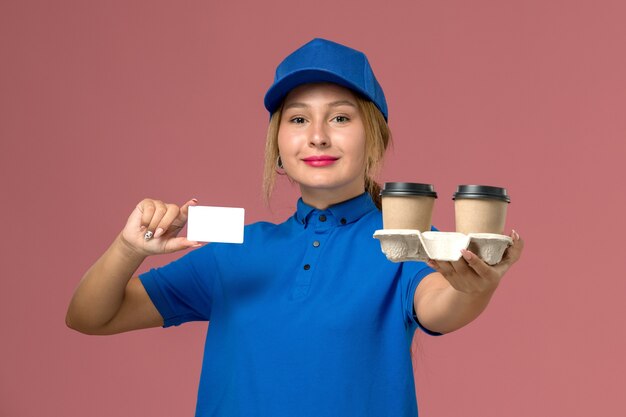 Mensajero femenino en uniforme azul sosteniendo tazas de entrega de café y tarjeta blanca en rosa, trabajador de entrega uniforme de servicio