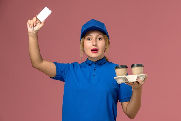 Mensajero femenino en uniforme azul sosteniendo tazas de entrega de café y tarjeta blanca en rosa, trabajo de entrega uniforme de servicio