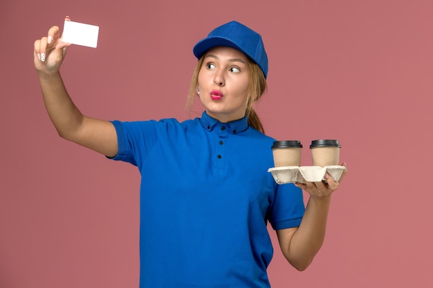 Mensajero femenino en uniforme azul sosteniendo tazas de entrega de café y tarjeta blanca en rosa, trabajo de trabajador de entrega uniforme de servicio