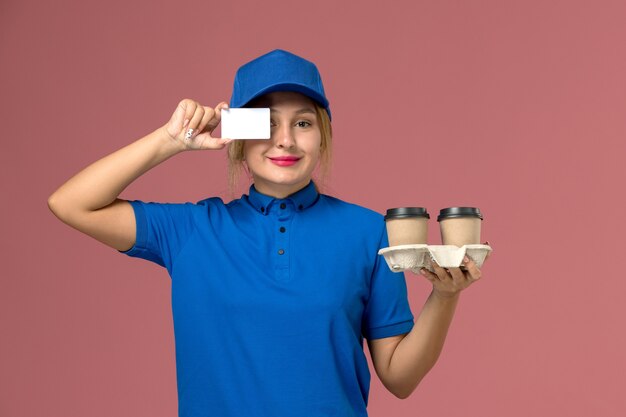 Mensajero femenino en uniforme azul sosteniendo tazas de entrega de café y tarjeta blanca con sonrisa en rosa, trabajo de trabajador de entrega uniforme de servicio