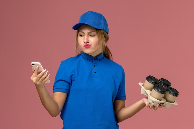 Mensajero femenino en uniforme azul sosteniendo el teléfono junto con tazas de café marrón en rosa claro, entrega uniforme del servicio de trabajo