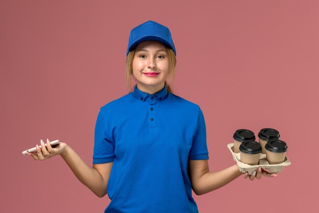 Mensajero femenino en uniforme azul sosteniendo el teléfono y con tazas de café marrón en rosa claro, trabajo de entrega uniforme de servicio