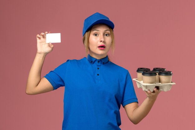 Mensajero femenino en uniforme azul con tarjeta blanca junto con tazas de café de entrega marrón en rosa claro, trabajo de servicio entrega uniforme