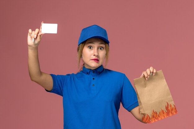 Mensajero femenino en uniforme azul con tarjeta blanca y paquete de alimentos en rosa claro, servicio de entrega uniforme de trabajo