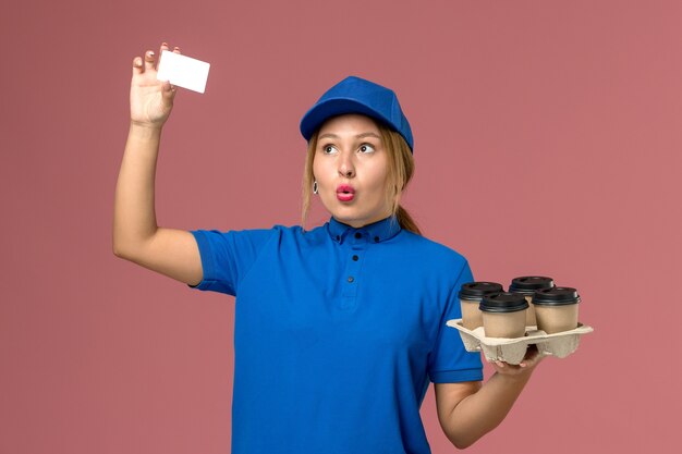 Mensajero femenino en uniforme azul con tarjeta blanca y tazas de café de entrega marrón en rosa claro, trabajador de entrega uniforme de trabajo de servicio